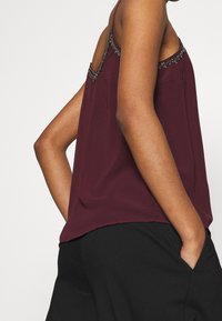 Personne portant un top sans manches bordeaux avec une garniture de perles décoratives et un pantalon noir, les mains dans les poches, devant un fond uni.