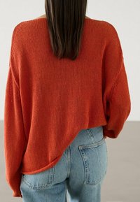 Luftig gestrickter, leuchtend orangefarbener Pullover mit weiten Ärmeln und asymmetrischem Saum, kombiniert mit hellblauen Jeans. Strukturierter Stoff und lässiges Design.