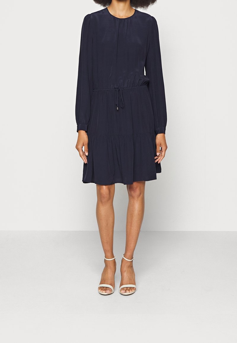 Femme portant une robe bleu marine longueur genou, manches longues, taille avec cordon de serrage, et des talons blancs ouverts avec bride à la cheville.
