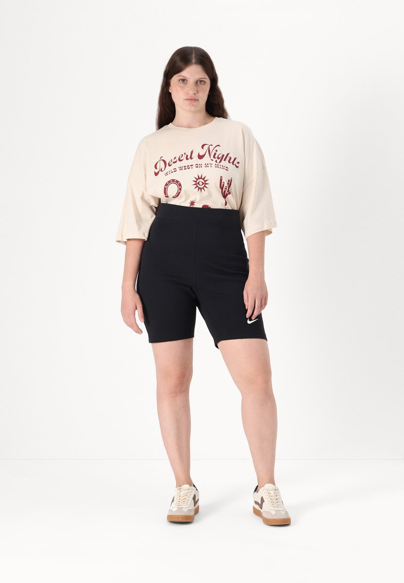 Vrouw in een beige oversized "Desert Nights" graphic t-shirt, zwarte Nike fietsshorts en beige sneakers, staand tegen een witte achtergrond.