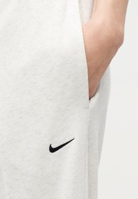 Χέρι μέσα στην τσέπη ανοιχτόχρωμων γκρι φόρμας Nike, με μικρό μαύρο λογότυπο Nike Swoosh κοντά στην τσέπη.