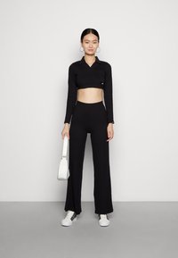 Top preto cropped de manga longa com gola, combinado com calças pretas de boca de sino. Acessórios incluem uma mala branca e sapatos com padrão.