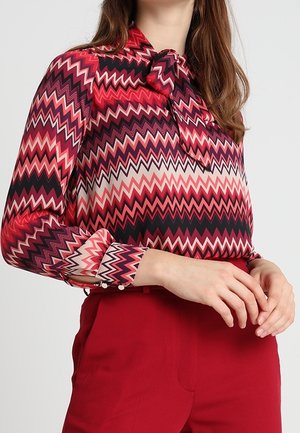 Vrouw draagt een blouse met lange mouwen en zigzagpatroon in rood, roze, zwart en wit met een dasspeld, gecombineerd met rood hooggetailleerde broek.