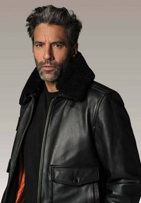 Homme avec des cheveux et une barbe poivre et sel portant une veste en cuir noire avec un grand col en fourrure et une chemise noire, fond neutre.