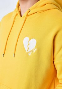 Sudadera amarilla hecha de tela suave, con un gráfico de corazón roto blanco en el pecho izquierdo y cordones a juego con puntas de metal.