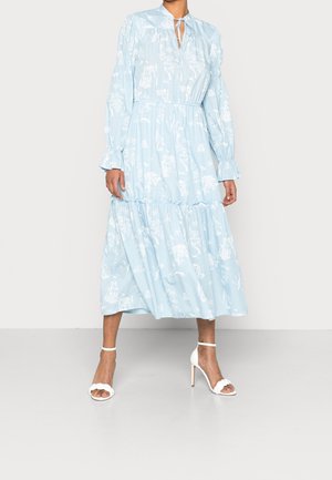 Vrouw draagt een lichtblauwe midi-jurk met lange mouwen en witte bloemenpatronen en witte sandalen met hoge hakken, staand op een effen achtergrond.