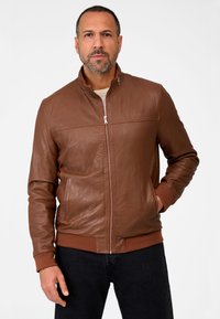 Bruine leren jas met een opstaande kraag, verticale gestikte details, geribbelde manchetten en een ritssluiting aan de voorkant. Draagt met donkere jeans.
