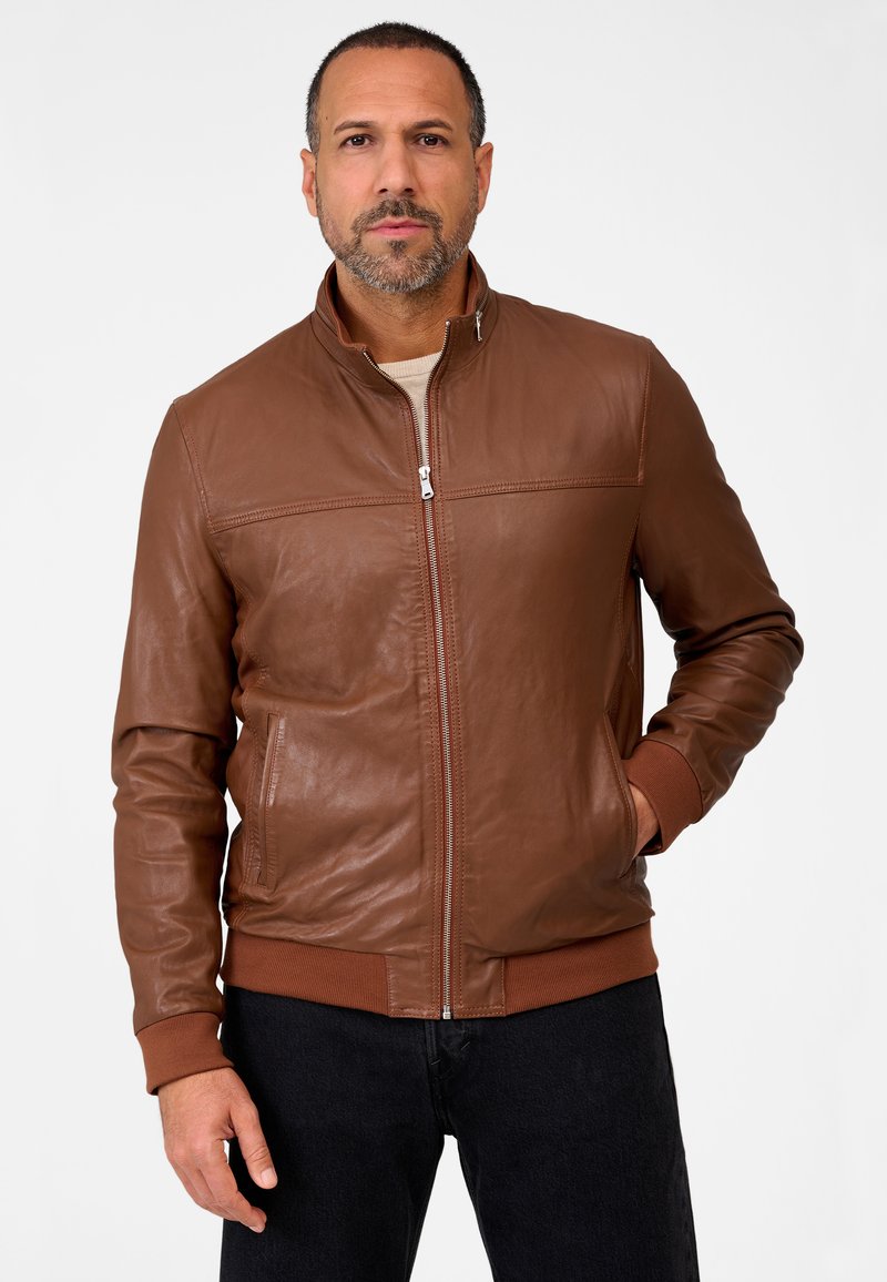 Brown leather jacket with a stand-up collar, vertical stitched detail, ribbed cuffs, and zippered front. Worn with dark jeans.