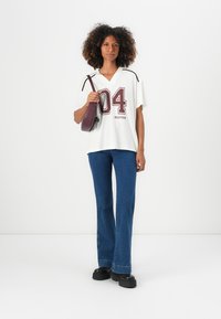 Weißes übergroßes T-Shirt mit maroonfarbener Zahl "04" und Text, kombiniert mit blauen Schlagjeans und einer kleinen lila Tasche. Schwarze Plattformschuhe.