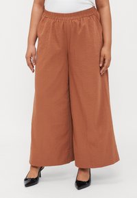 Pantalons larges marron réalisés en tissu texturé avec une ceinture élastique, associés à des escarpins à bout pointu noirs.