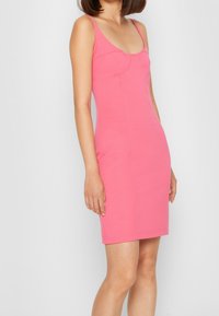 Dopasowana, bezrękawna sukienka w jasnoróżowym kolorze. Posiada dekolt w kształcie łódki oraz wyraźnie uformowane szwy, sięgająca powyżej kolana. Gładka tekstura.