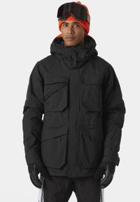 Chaqueta impermeable negra con múltiples bolsillos frontales, capucha ajustable y cremalleras. Lleva puesta una gorra de lana de color naranja brillante y guantes negros.