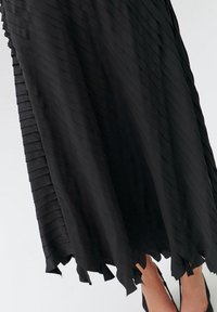 Sorte plisserede nederdel med et flydende design. Har struktureret stof og en asymmetrisk kant, der tilføjer dybde og bevægelse.