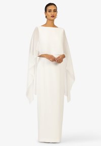 Long white dress with a straight silhouette, featuring a layered sheer cape draped over the shoulders and a boat neckline. Smooth texture.