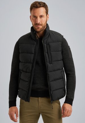 Gilet puffo nero con colletto alto, zip frontale, tasche laterali e maniche testurizzate. Indossato sopra un maglione di lana scuro.
