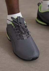 Närbild av en person som bär grå och neongröna Puma-sneakers med vita strumpor på ett slätt golv.