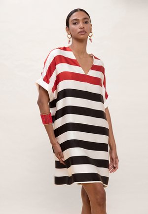 Mujer vestida con un vestido holgado con amplias franjas horizontales en rojo, negro y blanco, pendientes de aro dorados y una pulsera de cuentas rojas en un brazo.