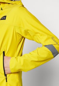 Keltainen vedenpitävä takki vetoketjullisella taskulla ja heijastavilla harmailla yksityiskohdilla hihoissa. Valmistettu hengittävästä materiaalista, jossa on huppu.