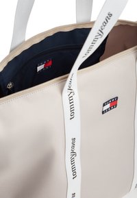 Μπεζ τσάντα ώμου με λευκές λαβές που φέρει τη λέξη "tommyjeans", εσωτερικό σε ναυτικό μπλε χρώμα και ένα μικρό λογότυπο με κόκκινες, λευκές και μπλε ρίγες.