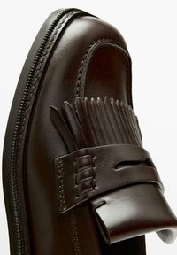Mocassin en cuir marron avec un détail à franges, surface lisse, forme étroite, et semelle noire contrastante avec des accents de couture.