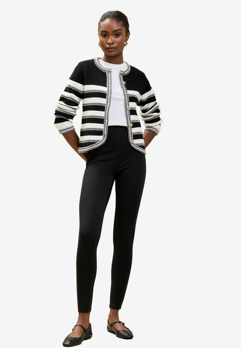 Schwarz-weiße gestreifte Strickjacke mit Rundhalsausschnitt, Knopfverschluss und strukturiertem Strick; kombiniert mit schwarzen Leggings und flachen Schuhen.