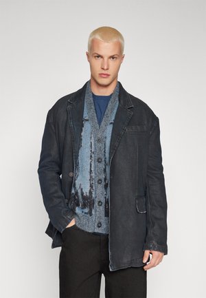 Mørkeblå denimjakke over en struktureret, flerfarvet blå cardigan med knapper, parret med en navy skjorte. Sorte bukser fuldender outfittet.