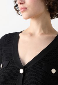 Haut en maille noir avec un décolleté en V, un tissu texturé et des accents de boutons argentés sur les poches poitrine. Col lisse et design ajusté.