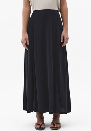 Personne portant une longue jupe noire et un haut beige, associée à des sandales plates noires, debout devant un fond blanc uni.