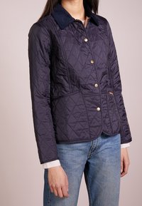 Marineblauwe gewatteerde jas met een corduroy kraag, gouden knopen en witte gestreepte manchetten. Kenmerkt zich door een getailleerd ontwerp en zijdelingse accentueringen.