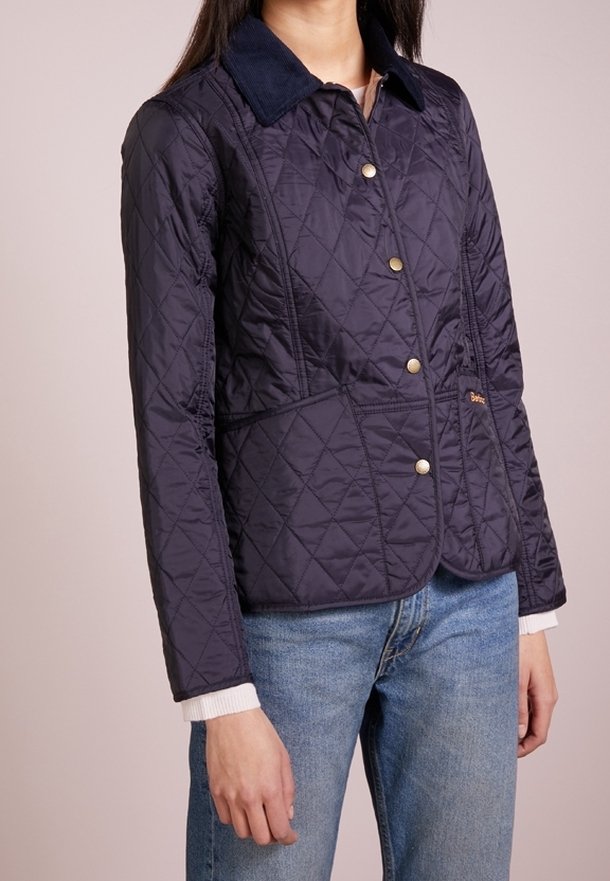 Marineblauwe gewatteerde jas met een corduroy kraag, gouden knopen en witte gestreepte manchetten. Kenmerkt zich door een getailleerd ontwerp en zijdelingse accentueringen.