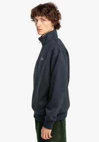 Young man with curly hair wearing a dark navy quarter-zip sweatshirt and green pants, standing in profile against a white background.