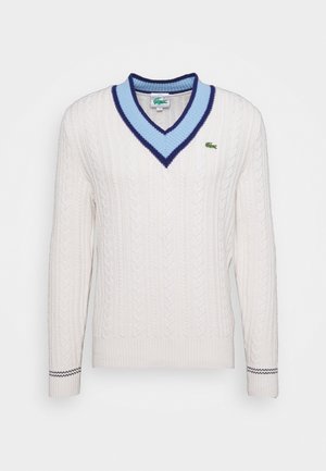 White cable-knit sweater with V-neck collar in light and dark blue, long sleeves, and small green crocodile logo on chest.