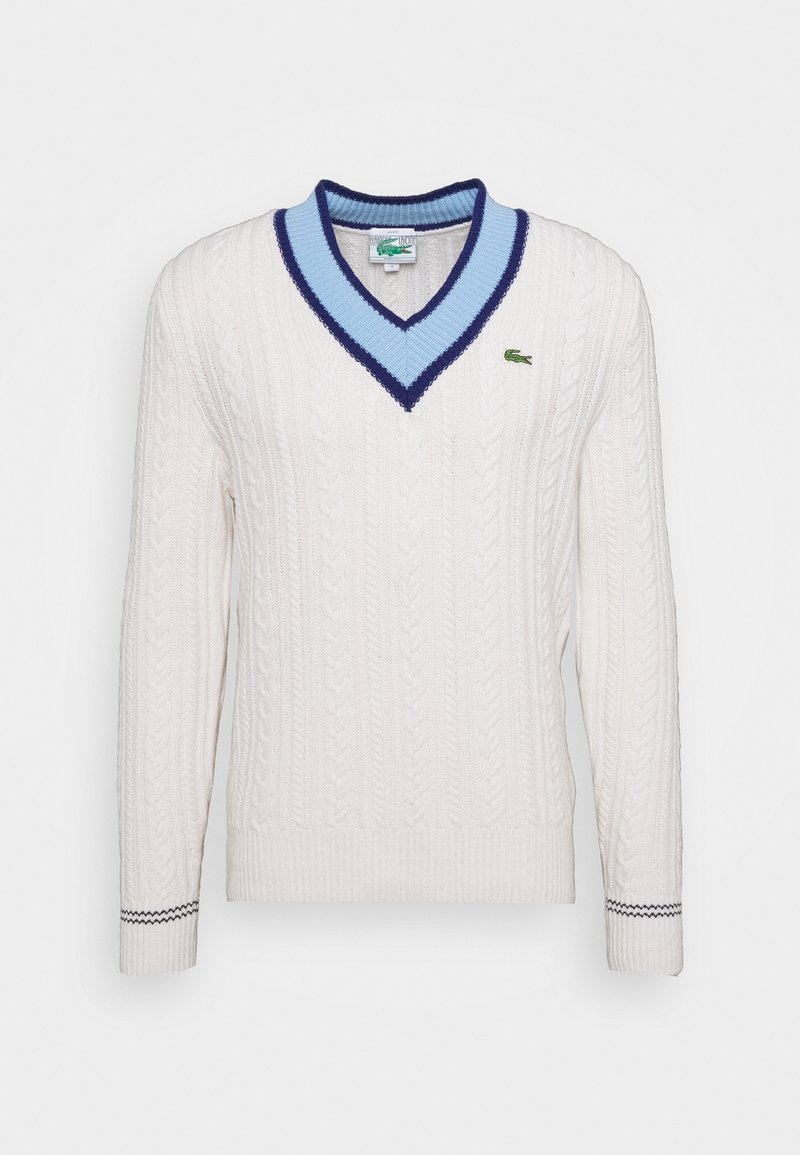 Pull en maille torsadée blanche avec col en V de couleur bleu clair et bleu foncé, manches longues, et petit logo crocodile vert sur la poitrine.