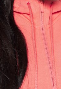 Felpa con zip di colore corallo, realizzata in tessuto testurizzato, con zip centrale e cappuccio. Accentuata da stringhe di regolazione su entrambi i lati.