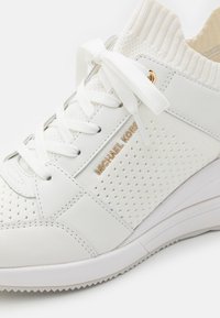 Baskets blanches Michael Kors avec col en tricot, panneau latéral perforé, lacets blancs noués en nœud et détail du nom de la marque en doré.
