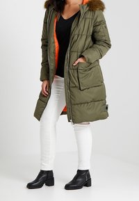 Olivgrüner gepolsterter Parka mit einer mit Pelz gefütterten Kapuze, Reißverschluss, seitlichen Taschen und einem orangefarbenen Innenfutter.