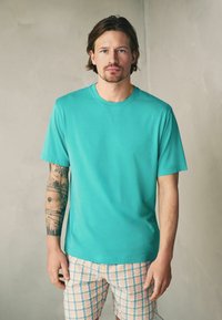 Homme aux cheveux mi-longs portant un t-shirt turquoise et un short à carreaux pastel, debout contre un mur beige uni.