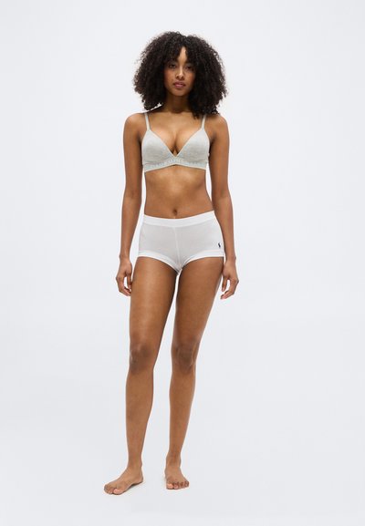 Γυναίκα με σγουρά μαλλιά, ξυπόλητη, φορώντας γκρι μπουστάκι και λευκά boxer briefs με λογότυπα μάρκας σε ένα απλό λευκό φόντο.