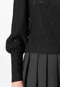 Top in maglia nera con un intricato motivo a diamante e maniche lunghe a sbuffo, abbinato a una gonna plissè grigio scuro.
