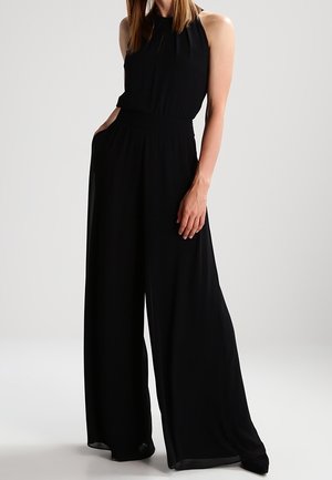 Combinaison noire à col halter en tissu léger, avec jambes larges, taille élastique et poches latérales. Texture lisse et fluide.