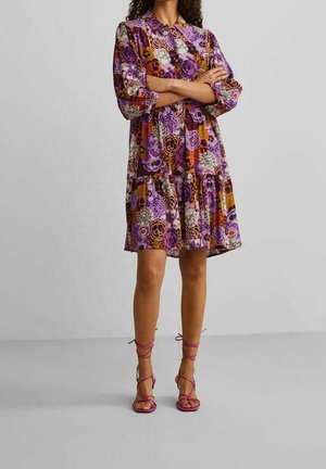 Bloemenjurk in paarse en oranje tinten, met kraag en pofmouwen, gelaagde rok, gecombineerd met roze sandalen met bandjes. Soepele stoftextuur.