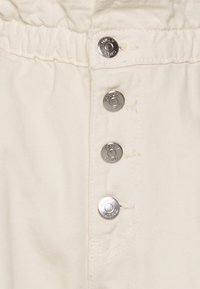 Vitt bomullstyg med tre glänsande silverknappar, sydda detaljer och ett elastiskt midjeband för justerbar passform.