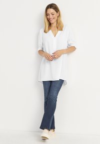 Valkoinen tunika V-pääntiellä ja taitetuilla lyhyillä hihoilla, yhdistetty tummansinisiin farkkuihin ja vaaleanbeigeihin korottomiin kenkiin. Silkkinen kangas ja rento istuvuus.