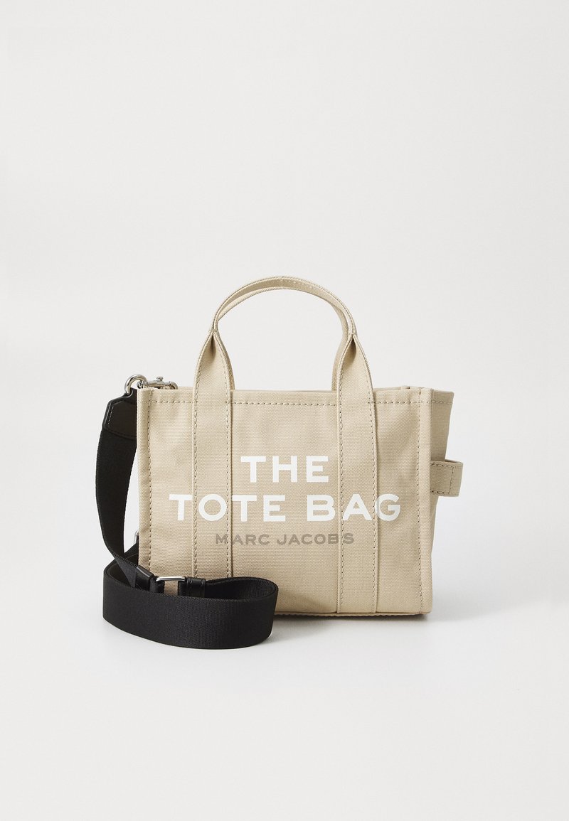 Beige canvas tote bag med dobbel top-håndtag, aftagelig sort strop og fed hvid tekst, der står "THE TOTE BAG MARC JACOBS."
