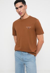 Καφέ βαμβακερό μπλουζάκι κοντό μανίκι, με στρογγυλή λαιμόκοψη και κεντημένο λογότυπο "HILFIGER" στο στήθος, συνδυασμένο με ανοιχτό μπλε τζιν.