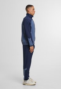 Navy krāsas trenera jaka un bikses ar gaiši zilu akcentu. Gluda auduma, ar rāvējslēdzēju kaklu, elastīgām piedurknēm un pieguļošs griezums. Valkāts kopā ar sporta apaviem.