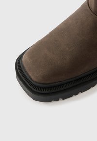 Barna bőr csizma négyzet alakú orral, texturált felülettel és fekete gumitalppal, amely bordázott széllel és sekély futófelületmintázatokkal rendelkezik.