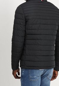 Svart puffer-jacka med horisontellt kviltade sektioner, huva och randig detalj vid kragen. Buren med blå jeans.