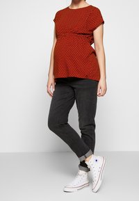 Rød graviditetsbluse med kort ærmer og prikker, samlet under brystet, parret med sorte stramme jeans og hvide højhælede sneakers.