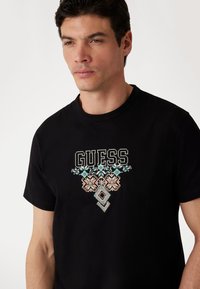 Must või aedviljadega kaunistus, millel on peene multivärviline broderie,  esiküljel on silmapaistev "GUESS" logo.
